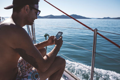 Side view of shirtless man using smart phone while sitting in boat on sea against clear sky during sunny day