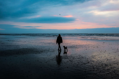 Silhouette mature woman with dog walking at beach against cloudy sky during sunset