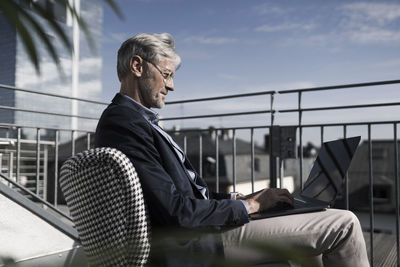 Grey-haired businessman sitting on balcony using laptop