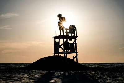 Silhouette man standing in lifeguard hut at beach against sky during sunset