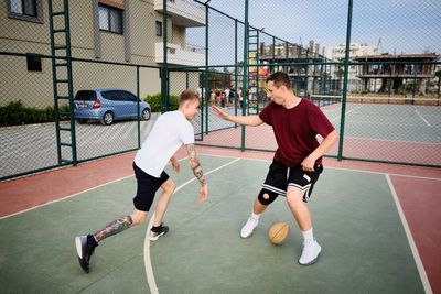 Two young adult men play basketball at outdoor court.