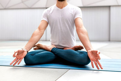 Cropped unrecognizable man in sportswear sitting in lotus pose and meditating with closed eyes and mudra gesture in spacious room with geometric wall and floor