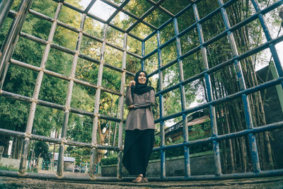 Low angle view of smiling woman leaning on metal grate against trees at park