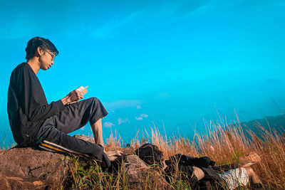 Side view of young man using smart phone against blue sky