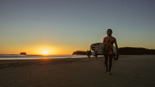 Shirtless man with surfboard walking at beach against sky during sunset