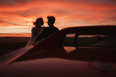 Silhouettes of bride and groom looking at each other while leaning on vehicle against cloudy sundown sky in bardenas reales natural park in navarra, spain