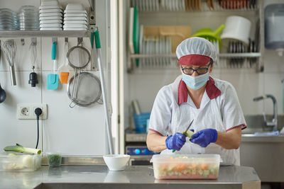 Female cook in white uniform and protective mask and gloves cutting fresh vegetables while preparing food in hospital kitchen during coronavirus pandemic