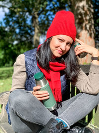 Young millennial woman in casual autumnal clothes siting on wooden bench in park and drinking coffee