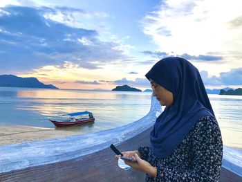 Side view of young woman using smart phone at beach against sky
