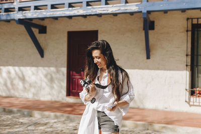 Young woman holding camera while standing against built structure