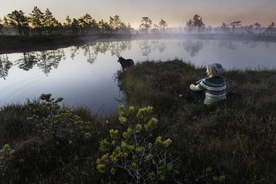 Woman and dog sit by pond at dawn with foggy atmosphere