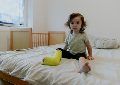 A baby girl with a cast on her leg sits on the bed.