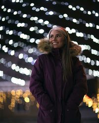 Low angle view of happy woman in warm clothing standing against illuminated lights at night
