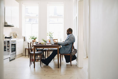 Full length of mature man working on laptop while sitting in kitchen at home