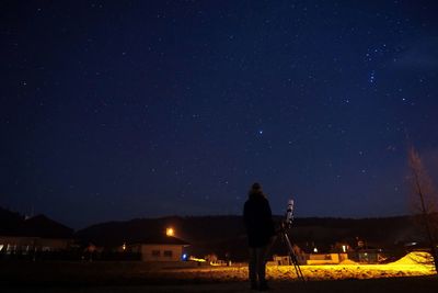 Rear view of man standing by telescope on field against sky at night