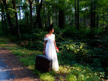 Woman holding luggage while standing on field in forest