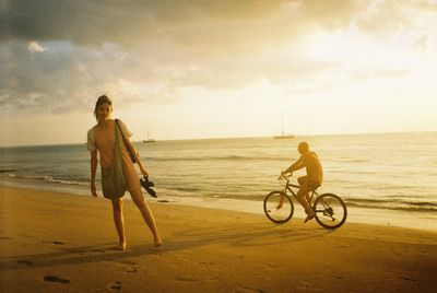 Full length portrait of beautiful woman standing at beach by man riding bicycle during sunset