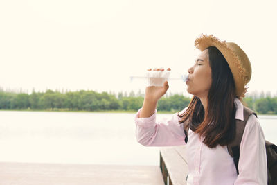 Thirsty young woman drinking water at lakeshore against clear sky