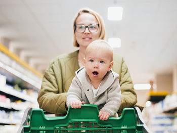 Mother pushing shopping cart with her infant baby boy child down department aisle in supermarket