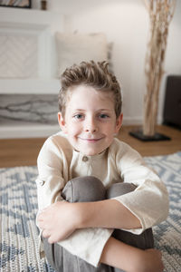 Cute boy in linen home clothes looks at a camera sitting on floor.