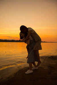 Woman piggybacking friend while standing at beach against sky during sunset