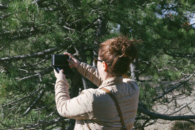 Rear view of woman photographing with smart phone by tree