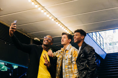 Positive diverse friends in casual clothes standing against staircase in underground passage and taking self portrait on smartphone