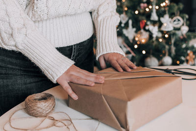 Midsection of woman wrapping christmas present in kraft paper in front of christmas tree at home