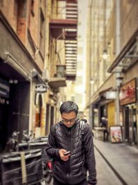 Portrait of young man using smartphone in alley against buildings in the city.