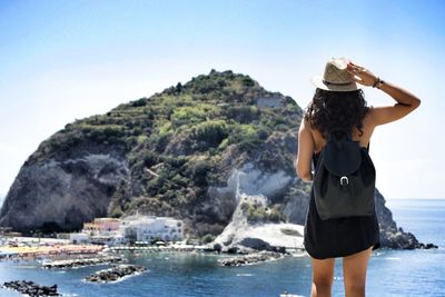 Rear view of young woman standing against mountain at beach