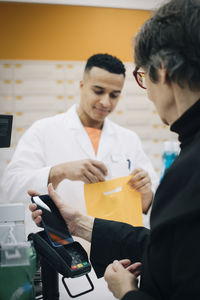 Senior female customer using contactless payment through mobile phone against pharmacist at checkout in store