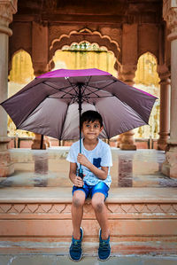 Full length of boy holding umbrella while sitting against historic building