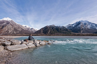 Man sitting on rock by lake against mountains