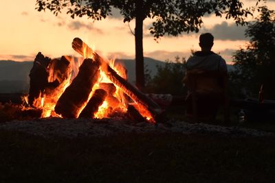 Rear view of silhouette man by campfire on field against sky at sunset