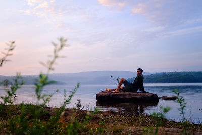 Man sitting on rock by lake against sky