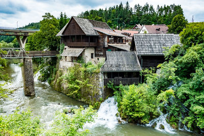 House by river amidst trees and houses against sky