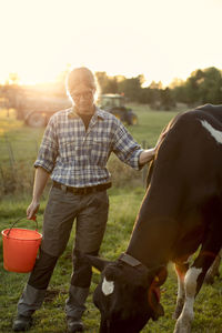 Female farmer standing with bucket by cow at field during sunset