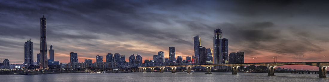 Illuminated buildings by river against sky during sunset