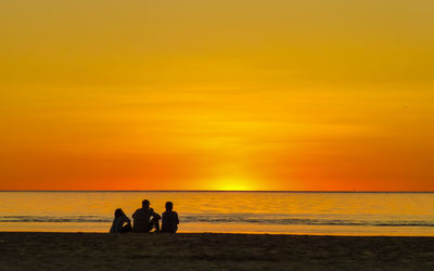 Silhouette people sitting at beach against orange sky