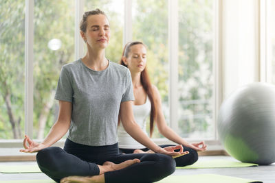 Woman practising yoga at the gym with the trainer sitting back to back on windows background