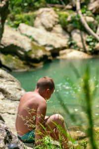 Rear view of shirtless boy on rock by lake