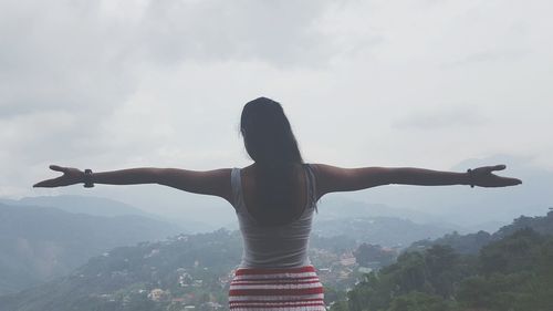 Rear view of woman standing with arms outstretched on mountain against cloudy sky
