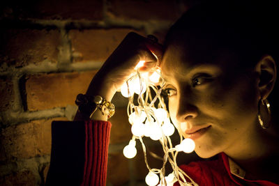 Close-up of woman holding illuminated lighting equipment while looking away by wall