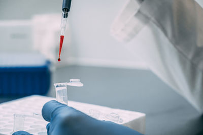 A scientist in blue gloves gently squeezes a red liquid from a pipette into a container. 