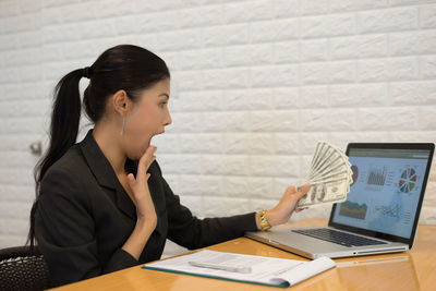 Shocked businesswoman holding paper currency over laptop at desk in office