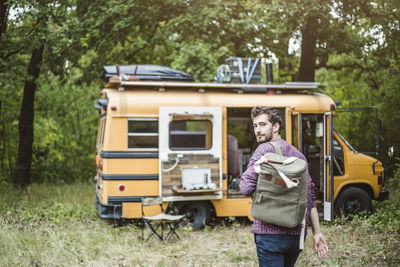 Rear view portrait of young man walking with backpack towards motor home in forest during camping