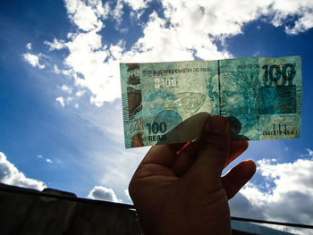 Cropped hand holding paper currency against sky