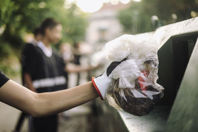 Cropped hand of teenage boy holding plastic garbage bag by recycling bin