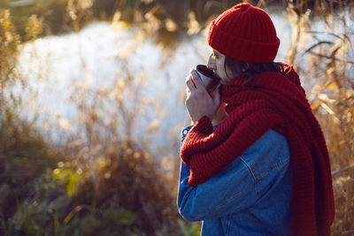 Man in a red knitted scarf and hat stands by the lake in autumn and drinks tea mug thermos