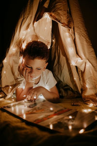 Blondy preschooler boy reads a book in hut at home with garlands and lights at night